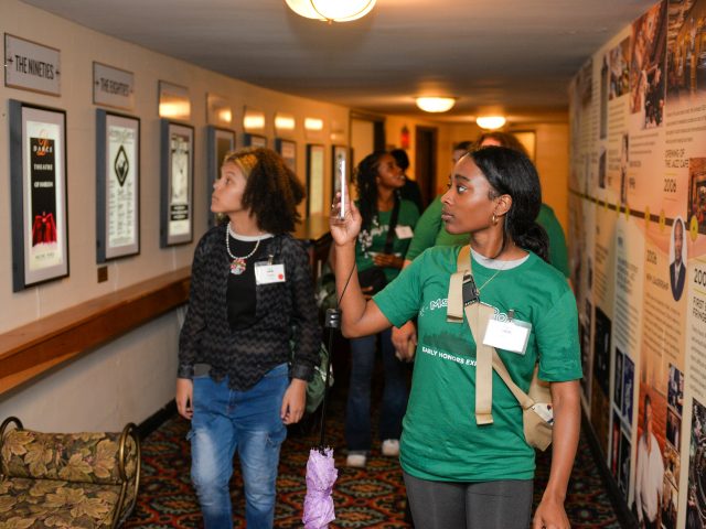 Students walk down a hallway where the walls are covered with music history information. One student holds out her phone to take a photo.