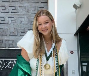 Destiny Kanning smiling in a graduation robe, cords, and medal. In the background is the MSU Athletics area in 1855 Place.